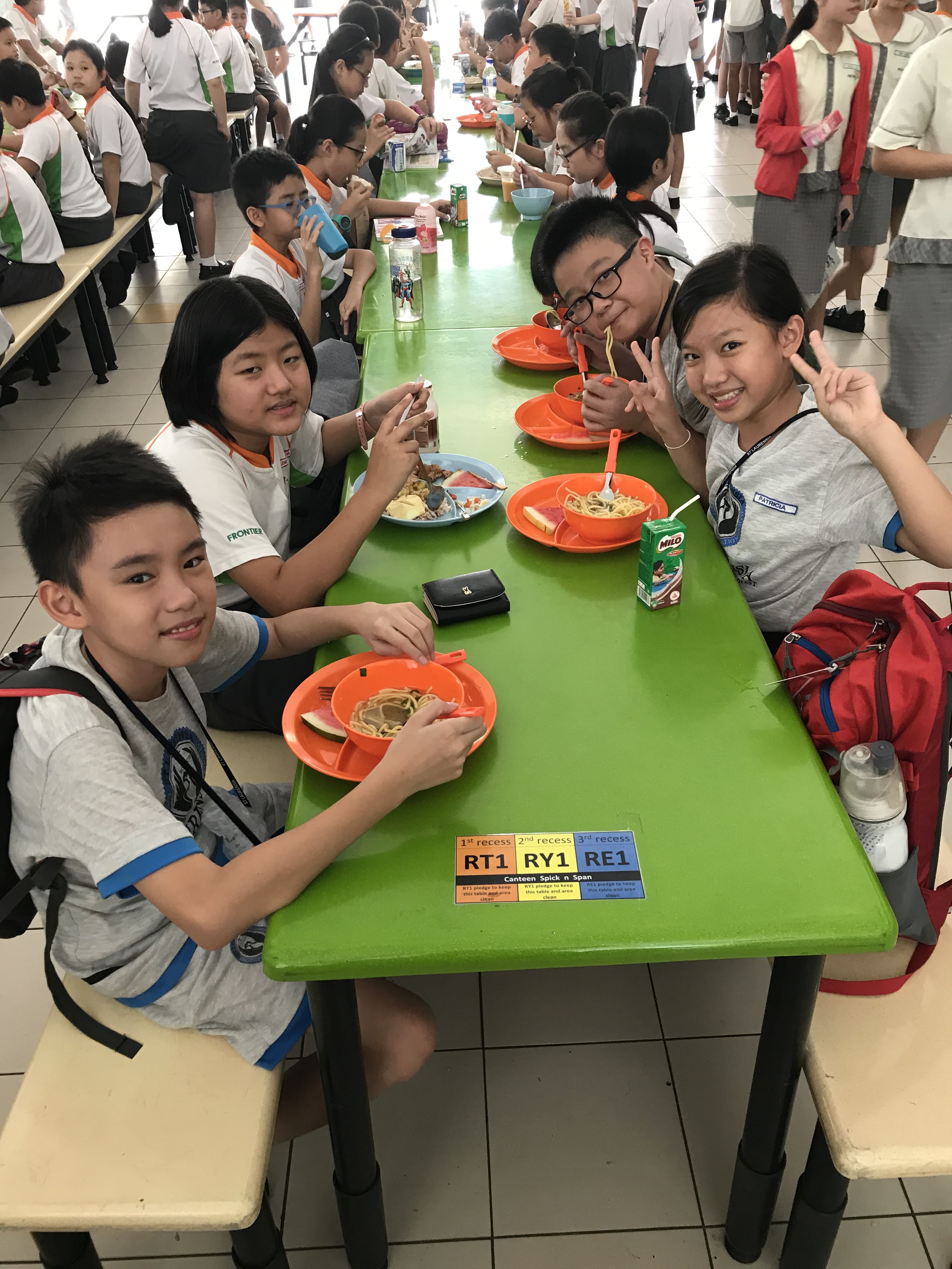 Four school children eating noodles on orange trays at a green table in a busy school canteen.